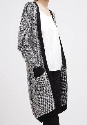 Robe cardigan longue avec un motif tricoté noir et blanc. Présente une ouverture sur le devant, des poches latérales et des poignets côtelés. Tissu texturé et coupe décontractée.