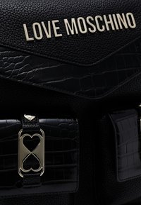 Svart läderhandväska med krokodilåtergivning och guldfärgad detaljer. Har "LOVE MOSCHINO"-logotypen och hjärtformade mönster.