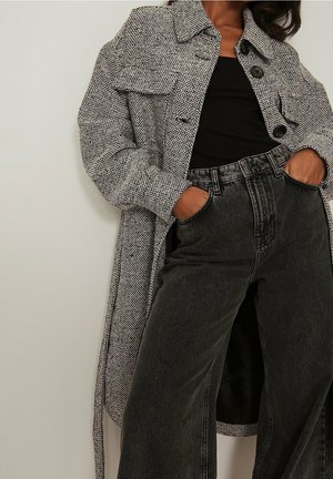 Femme portant un long manteau gris texturé, un haut noir côtelé et un jean noir délavé taille haute, les mains dans les poches, sur fond clair.