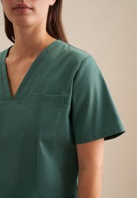 Top a manica corta con scollo a V in tessuto verde scuro uniforme. Presenta una tasca a toppa sul lato sinistro del petto e una texture liscia e opaca.