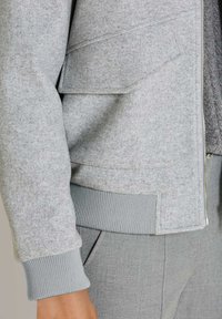 Nahaufnahme einer Person, die eine hellgraue Wolljacke mit gerippten Bündchen und Saum trägt, kombiniert mit passenden grauen Hosen.