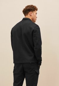 Jachetă neagră cu o textură netedă și o croială ajustată, având un guler și mâneci lungi, asortată cu pantaloni corespunzători.