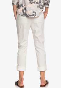 Valkoiset pellavapohkeiseen asti ulottuvat housut, joissa on käännetyt lahkeet, sileä rakenne, rento istuvuus ja jotka on yhdistetty sandaalien kanssa. Takakuvassa näkyy kaksi taskua.