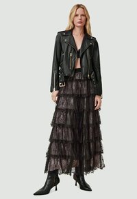 Femme blonde portant une veste de motard en cuir noir, une jupe maxi en dentelle noire à franges, un bralette noir et des bottes chelsea noires à bout pointu.