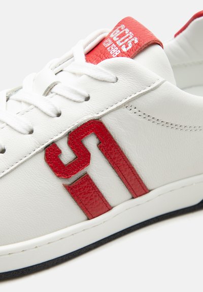 Λευκό δερμάτινο αθλητικό παπούτσι με λευκά κορδόνια, κόκκινες ρίγες λογότυπου στο πλάι και κόκκινη ετικέτα στη γλώσσα με ανάγλυφο κείμενο της μάρκας.