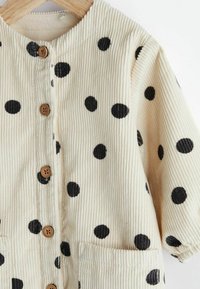 Beige corduroyjas met zwart stippenpatroon, ronde halslijn, sluiting met knopen aan de voorkant en twee zakken aan de voorkant.