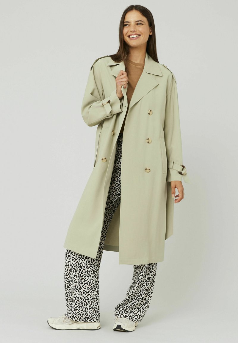 Femme souriante portant un trench-coat beige, un haut marron, un pantalon à motifs noir et blanc et des baskets blanches, se tenant devant un fond uni.