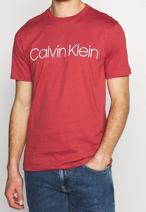 Mężczyzna w czerwonej koszulce z krótkim rękawem z białym logo „Calvin Klein” na klatce piersiowej i niebieskich dżinsach, ręka w kieszeni.