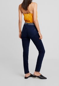 Los jeans skinny azul oscuro presentan una cinturilla texturizada con un patrón, combinados con una blusa amarilla de satén y zapatos negros tipo slip-on.