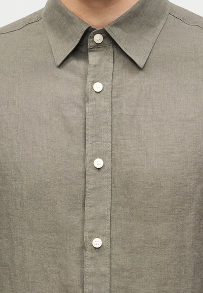Άνδρας που φοράει μια μονόχρωμη, κουμπωτή λινή πουκάμισο ελιάς με μυτερό γιακά.