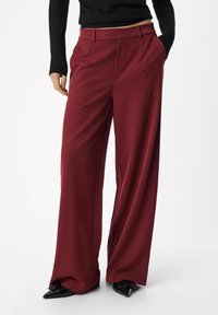 Bordeaux wijde broek gemaakt van gladde stof, met een hoge taille, voorplooien en schuine zakken. Combinatie met zwarte hakken.