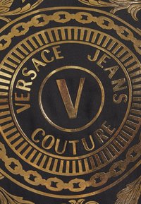 Musta kangas rohkealla kultaisella tekstillä, jossa lukee "VERSACE JEANS COUTURE" ja keskellä on "V" -symboli, jota ympäröi monimutkaiset kuviot.