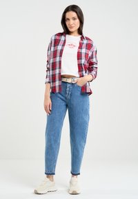 Karierter Hemd in Rot und Blau über einem weißen, verkürzten T-Shirt, kombiniert mit hellblauen, hochgeschnittenen Jeans und beigen Sneakers.