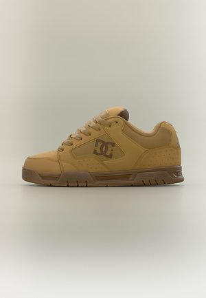 Niski sneaker marki Tan DC w kolorze brązowym z wyściełanym kołnierzem, teksturowanym panelem bocznym i grubą gumową podeszwą, pokazany z boku na jednolitym tle.