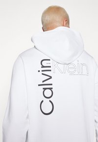 Felpa bianca con cappuccio caratterizzata da un grande logo nero "Calvin Klein" stampato verticalmente sul retro, con il dettaglio "Est. 1968" sopra.