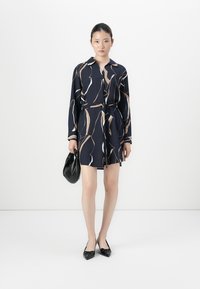 Femme portant une robe longue à manches longues à motifs navy, ceinturée, tenant un sac à main noir et portant des ballerines noires à bouts pointus, se tenant contre un fond blanc.