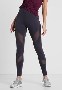 Legginsy z wysokim stanem w ciemnoszarym materiale z przezroczystymi panelami siateczkowymi w wzór chevron. W zestawieniu z szarymi butami sportowymi.