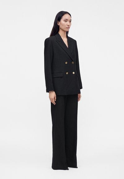 Femme debout portant un blazer noir à double boutonnage avec des boutons dorés et un pantalon large assorti sur un fond uni.