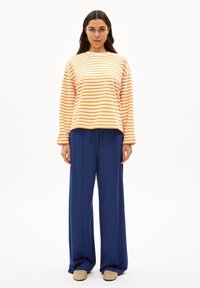 Top a righe giallo e arancione con maniche lunghe, abbinato a pantaloni a gamba larga neri a vita alta. Tessuto morbido con vestibilità rilassata. Scarpe beige senza lacci.