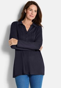 Navyblaues Langarmhemd mit Kragen, Knopfleiste und gefälteltem Vorderdesign. Kombiniert mit hellblauen Jeans.