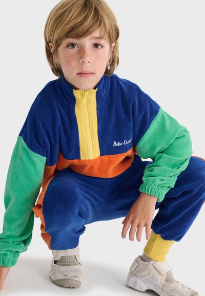 Enfant aux cheveux châtain clair portant un survêtement à blocs de couleur bleu, vert, orange et jaune, accroupi, regardant droit devant sur un fond uni.