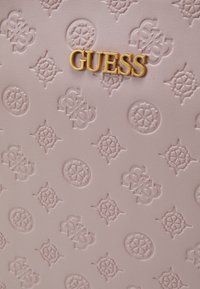 Simili cuir rose clair avec des motifs en relief, portant en évidence un logo "GUESS" doré. La surface texturée ajoute du détail.