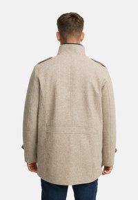 Manteau en laine beige avec un col montant, des épaulettes et une fente au dos. Il présente une texture lisse et une coupe ajustée, porté avec un jean foncé.