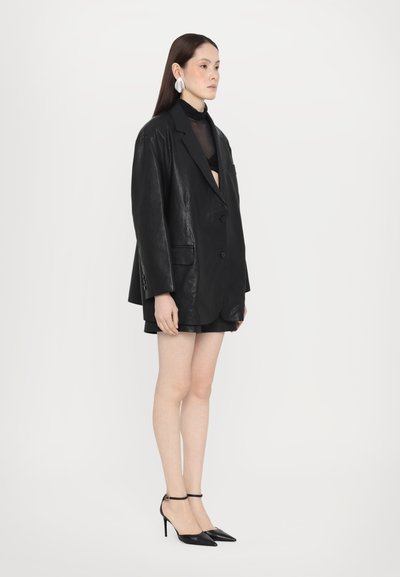 Sacou din piele neagră supradimensionat, cu revere înnodat, asociat cu un top transparent negru și pantaloni scurți negri, completat de tocuri negre ascuțite.