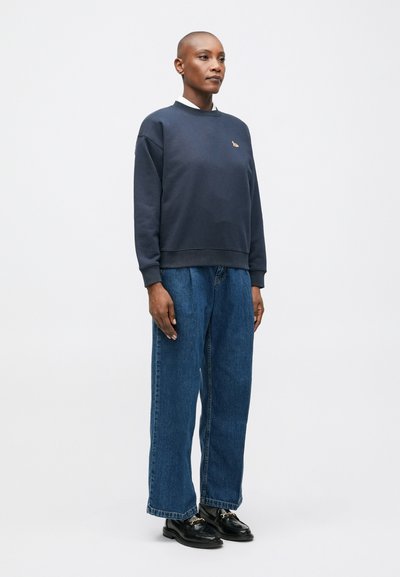 Jūras spēku džemperis ar ribinātu apkakli, pārmērīgs piegriezums, apvienots ar platām zils biksēm un melnām zandēm. Diskreta logo akcentācija uz krūtīm.