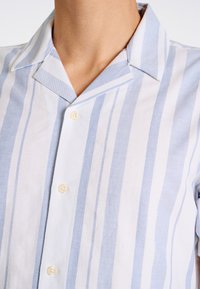 Camicia a maniche corte azzurro chiaro e bianca a righe con colletto da campeggio, caratterizzata da una chiusura con bottoni e tessuto testurizzato. I bottoni sono color crema.