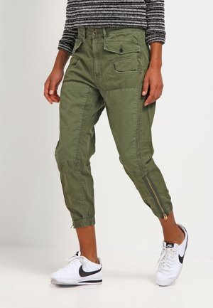 Olivengrønne cargobukser med elastiske manchetter, flere lommer og lynlåsdøre i siden, kombineret med hvide og sorte sneakers.