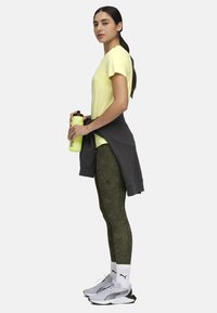 Camiseta deportiva de color amarillo claro, sudadera gris atada a la cintura, leggings de oliva con estampado, zapatillas blancas con detalles negros, sosteniendo una botella de agua verde.