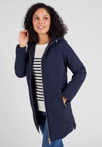 Chaqueta acolchada de color azul marino con capucha, cremallera dorada y bolsillos laterales, combinada con una camiseta de rayas y vaqueros azul. Textura suave y diseño moderno.