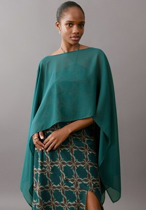 Donna che indossa uno scialle trasparente color verde acqua sopra un vestito stampato verde acqua con spacco laterale, in piedi davanti a uno sfondo grigio uniforme.