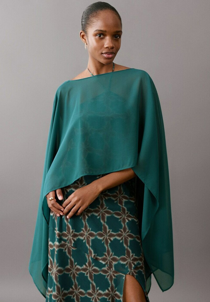 Frau trägt tealfarbene transparente Stola über einem gemusterten tealfarbenen Kleid mit Seitenschlitz, steht vor schlichtem grauem Hintergrund.