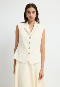 Ærmeløs cremefarvet vest med skræddersyet pasform, der har guldknapper og to forlommer, fremstillet af glat stof for et struktureret udseende.