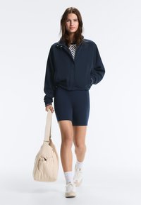 Nainen kävelee yllään tummansininen takki, sitä vastaavat pyöräilyshortsit, valkoiset lenkkarit ja suuri beige olkalaukku.