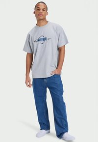 T-shirt cinza de algodão com mangas curtas e um logótipo gráfico. Combinada com calças de ganga de corte largo em azul claro, usadas sobre meias brancas.