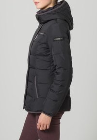 Žena v černé zateplené bundě s kapucí, se zipovými kapsami na hrudi a bocích, v kombinaci s vínovými kalhotami, zachycena z boku.