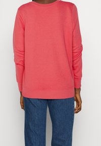 Sudadera coral de mangas largas, con cuello redondo y puños acanalados. Textura suave, combinada con jeans rectos de tela vaquera azul oscuro.