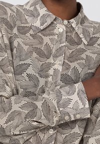 Camicia beige con bottoni, caratterizzata da un motivo astratto nero, colletto a punta e bottoni simili a perle, realizzata in un tessuto liscio.