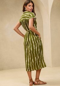 Robe midi rayée verte et blanche avec des manches courtes, taille cintrée, poches et ceinture assortie. Des sandales plates sont portées.