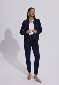 Granatowa kurtka bombowa z złotym zamkiem, biała bluzka w serek oraz dopasowane granatowe spodnie slim-fit. Jasnobeżowe szpilki ze złotymi akcentami dopełniają stylizację.