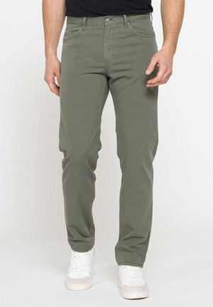 Uomo con pantaloni verdi oliva a taglio dritto e sneakers bianchi in piedi davanti a uno sfondo bianco uniforme.