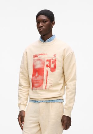 Béžová mikina s červeným grafickým potlačou zobrazujúcou tvár a fotoaparát, s rebrovanými manžetami a lemom, nosená cez svetlomodrú rifľovú košeľu.