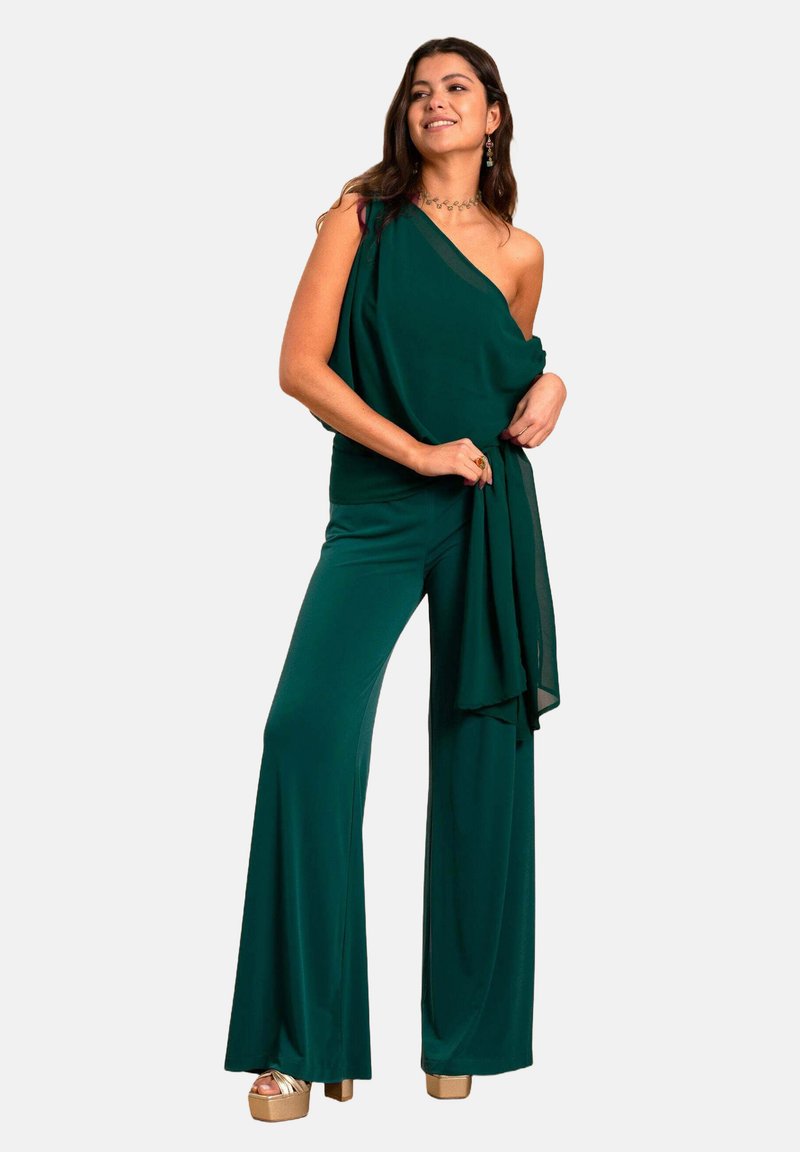Groene off-shoulder jumpsuit van lichtgewicht stof, met wijde benen en een vloeiende strikdetail bij de taille. Gouden plateau schoenen maken de look compleet.