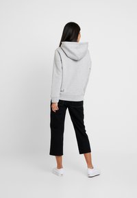 Kobieta stojąca plecami, ubrana w jasnoszary hoodie, czarne spodnie 7/8 i białe trampki, na tle jednolitego białego tła.