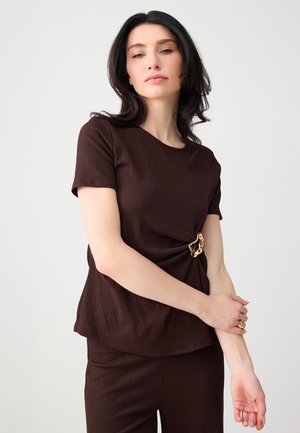 Femme aux cheveux bruns ondulés portant un haut marron foncé texturé à manches courtes avec une boucle décorative dorée et un pantalon assorti.