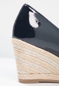 Námořnické patentové klínové podpatky s pleteným jutovým základem, zdobené kovovými pruhy. Design s špičatou špičkou. Hladký, lesklý povrch.