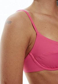 Rosa Bikini-Oberteil mit dünnen Trägern, nahtlosem Design und moderater Bedeckung. Hauttextur auf dem Oberarm sichtbar, mit einem Tattoo, das "BABY" liest.